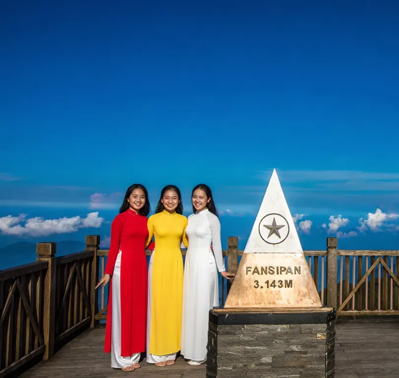 Three young women in traditional Vietnamese ao dai posing beside the Fansipan 3,143m summit marker in Sapa